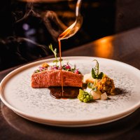Ein kunstvoll angerichtetes Gourmetgericht mit Fleisch, Gemüse und Sauce auf einem eleganten Teller. Eine Sauce wird darüber gegossen., © wosilatfotografie