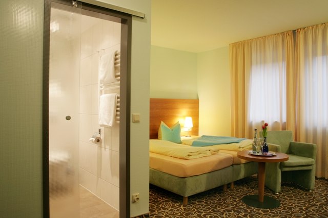 A cozy double room with a bed, table, chairs, and an adjoining bathroom. Light-colored curtains and carpet create a welcoming atmosphere., © ateckhotel A cozy double room with a bed, table, chairs, and an adjoining bathroom. Light-colored curtains and carpet create a welcoming atmosphere., © ateckhotel