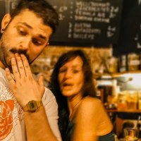 Drei Personen in einer Bar, eine mit Hand vor dem Mund, eine l&auml;chelnd, eine mit M&uuml;tze. Im Hintergrund eine Getr&auml;nketafel., &copy; Caf&eacute; Galao