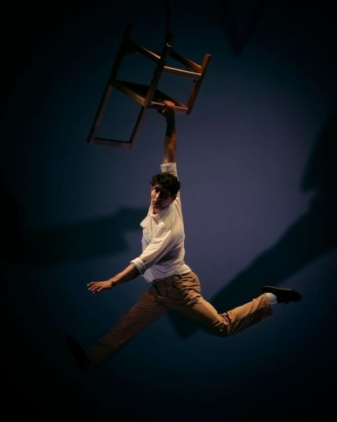 Ein Mann in einem wei&szlig;en Hemd und beigen Hosen h&auml;ngt an einem schwebenden Stuhl in einem dunklen Raum. Die Szene wirkt dramatisch und dynamisch., &copy; Theaterhaus Stuttgart e.V.
