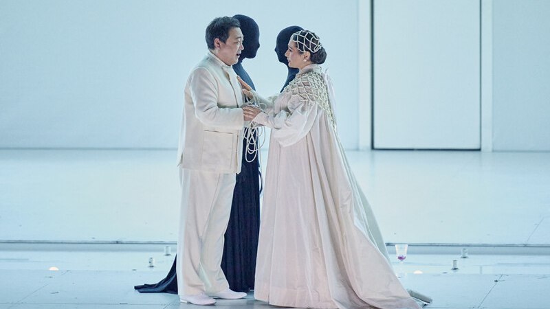 Zwei Personen in weißen Kostümen stehen sich gegenüber, begleitet von zwei schwarz gekleideten Figuren auf einer Bühne., © Württembergische Staatstheater Stuttgart Zwei Personen in weißen Kostümen stehen sich gegenüber, begleitet von zwei schwarz gekleideten Figuren auf einer Bühne., © Württembergische Staatstheater Stuttgart