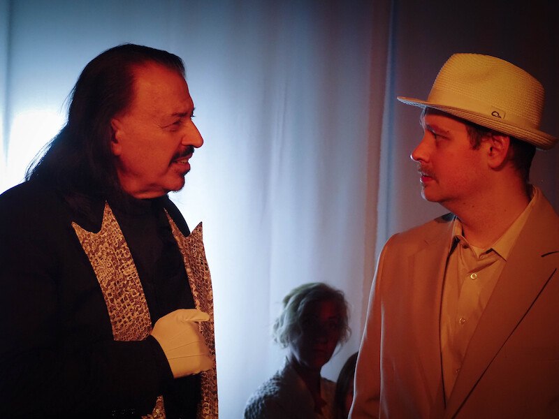 Sieben Personen in wei&szlig;er Kleidung posieren vor neutralem Hintergrund. Eine Person h&auml;lt einen Hut. Alle blicken in verschiedene Richtungen., &copy; Theater Atelier