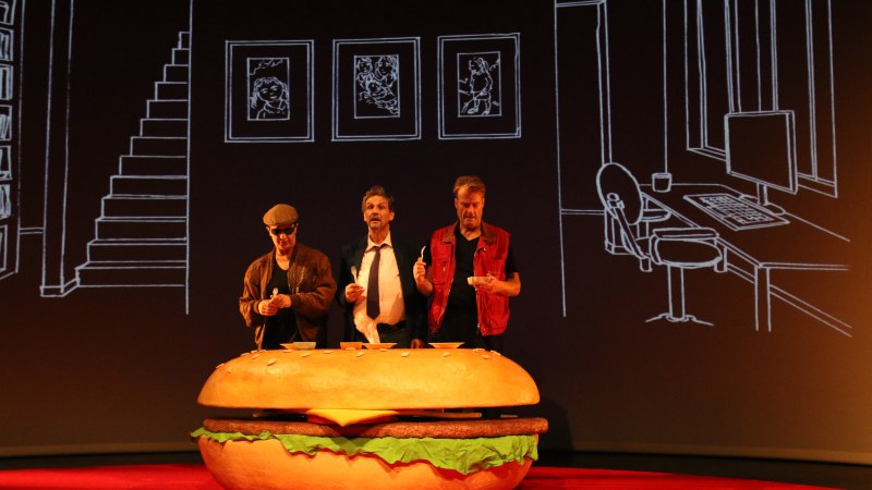 Drei M&auml;nner stehen hinter einem riesigen Hamburger-Requisit auf einer B&uuml;hne. Der Hintergrund zeigt gezeichnete M&ouml;bel und Bilderrahmen., &copy; Sabine Haymann