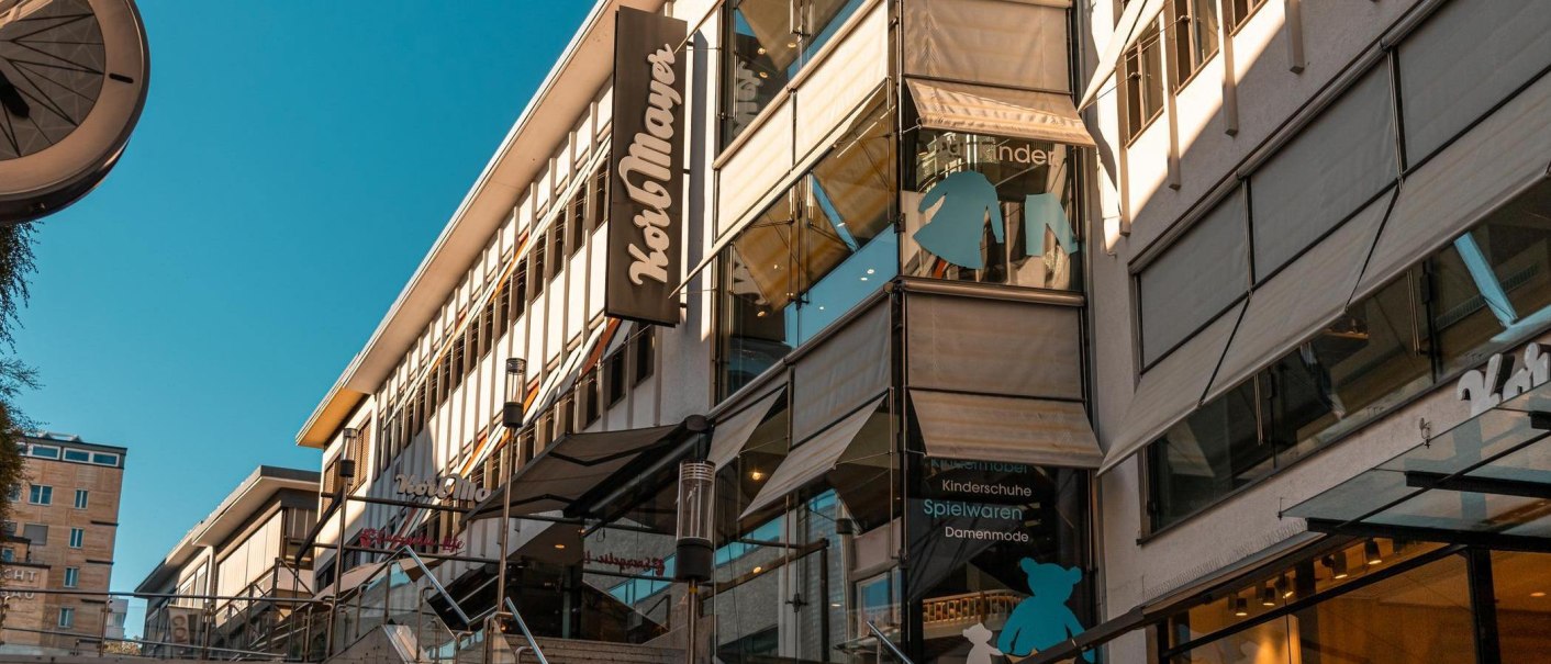 Gebäude mit Korbmayer-Schild, Schaufenster mit Kinderkleidung und Spielwaren. Moderne Architektur unter blauem Himmel., © Stuttgart Marketing GmbH, Sarah Schmid