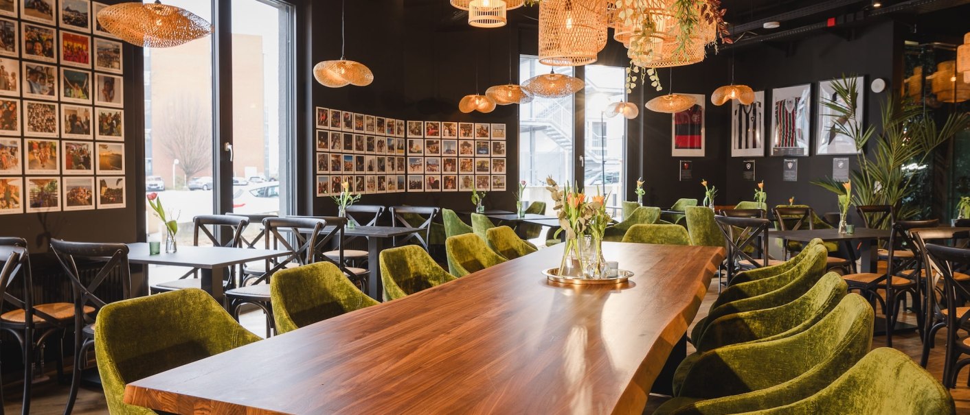 Ein Café mit grünen Samtstühlen, Holztischen und dekorativen Hängelampen. An den Wänden hängen viele Fotos., © TOMAS