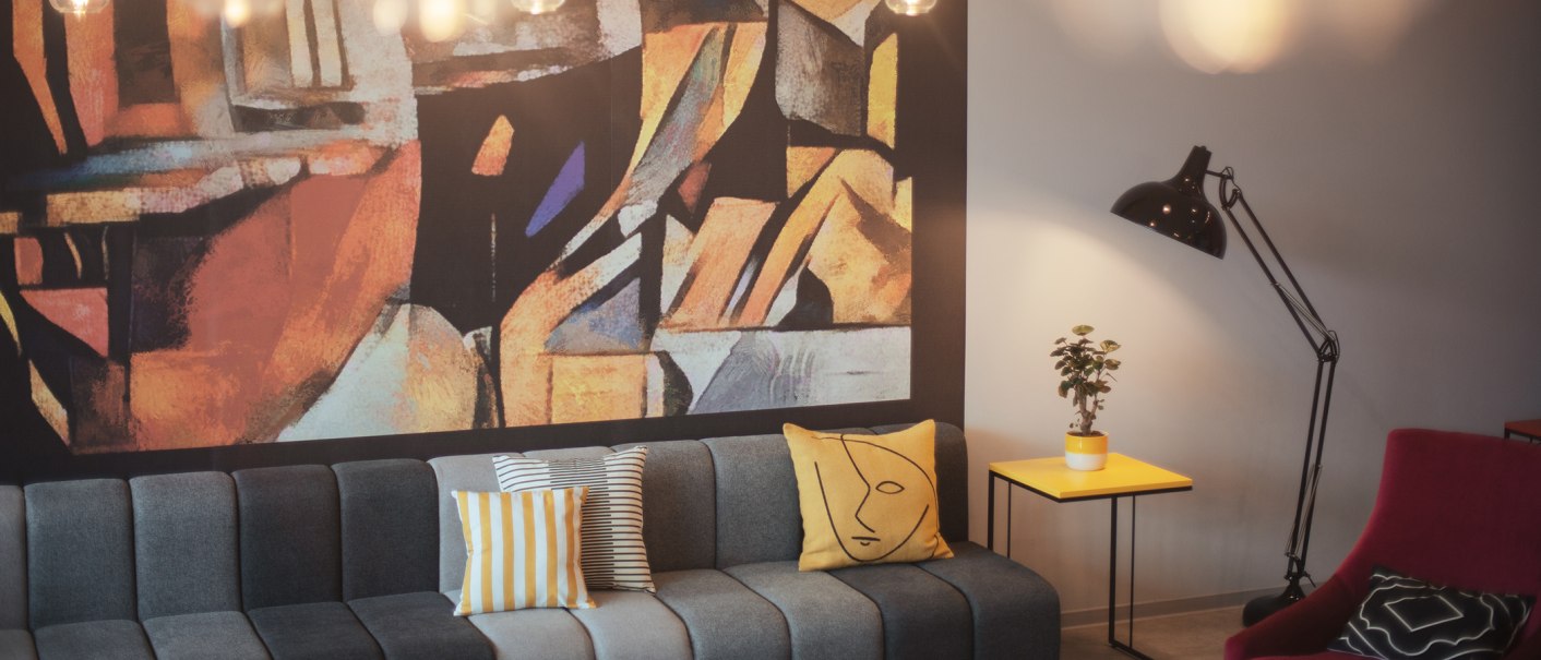 A modern lobby with a gray sofa, colorful pillows, an abstract wall hanging, a yellow side table, a plant, and a floor lamp., © the niu Form - Novum Hospitality A modern lobby with a gray sofa, colorful pillows, an abstract wall hanging, a yellow side table, a plant, and a floor lamp., © the niu Form - Novum Hospitality