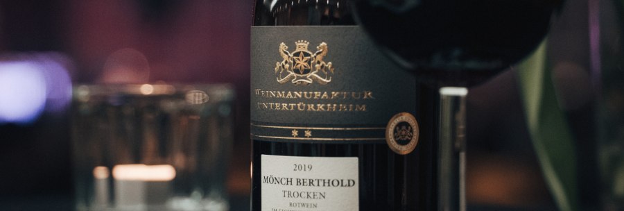 Close-up of a "Mönch Berthold" wine bottle from 2019, dry, with a glass of red wine next to it. Elegant label with gold lettering., © Weinmanufaktur Untertürkheim