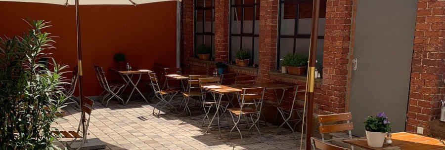 Cozy courtyard with wooden tables and chairs, parasol and plants. Red brick walls and paved floor create an inviting atmosphere., &copy; Gustav Gastro GmbH