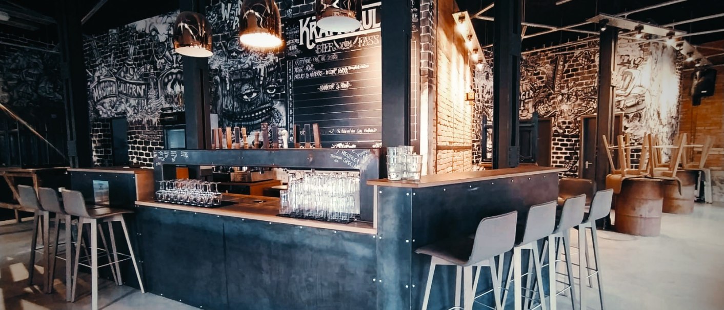 Moderne Bar mit industriellem Design, hohen Stühlen und stilvollen Lampen. Wände mit Graffiti und eine Tafel mit Getränkenamen., © Kraftpaule GmbH