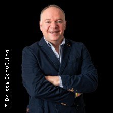 Ein Mann in einem dunklen Anzug lächelt mit verschränkten Armen vor einem schwarzen Hintergrund., © links im Bild Ein Mann in einem dunklen Anzug lächelt mit verschränkten Armen vor einem schwarzen Hintergrund., © links im Bild