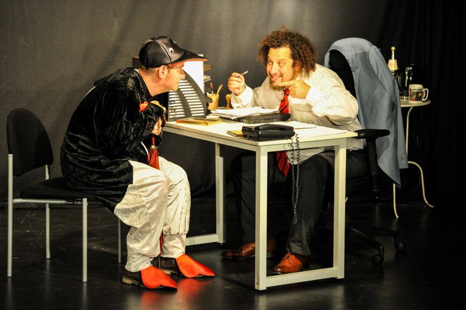 Zwei Männer in einem Büro, einer im Anzug, der andere in einem Kostüm, sitzen sich an einem Schreibtisch gegenüber und diskutieren lebhaft., © Theater am Olgaeck / C. Schöch