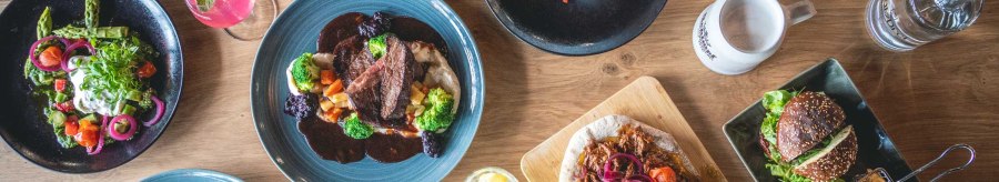 Ein Tisch mit verschiedenen Gerichten: Fleisch mit Gemüse, Salate, Burger mit Pommes, Brot und Desserts. Getränke sind ebenfalls zu sehen., © SatzDrei