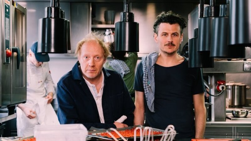 Zwei Männer in einer modernen Restaurantküche, umgeben von Küchenutensilien und Lebensmitteln. Im Hintergrund arbeitet ein Koch., © Theaterhaus Stuttgart e.V. Zwei Männer in einer modernen Restaurantküche, umgeben von Küchenutensilien und Lebensmitteln. Im Hintergrund arbeitet ein Koch., © Theaterhaus Stuttgart e.V.