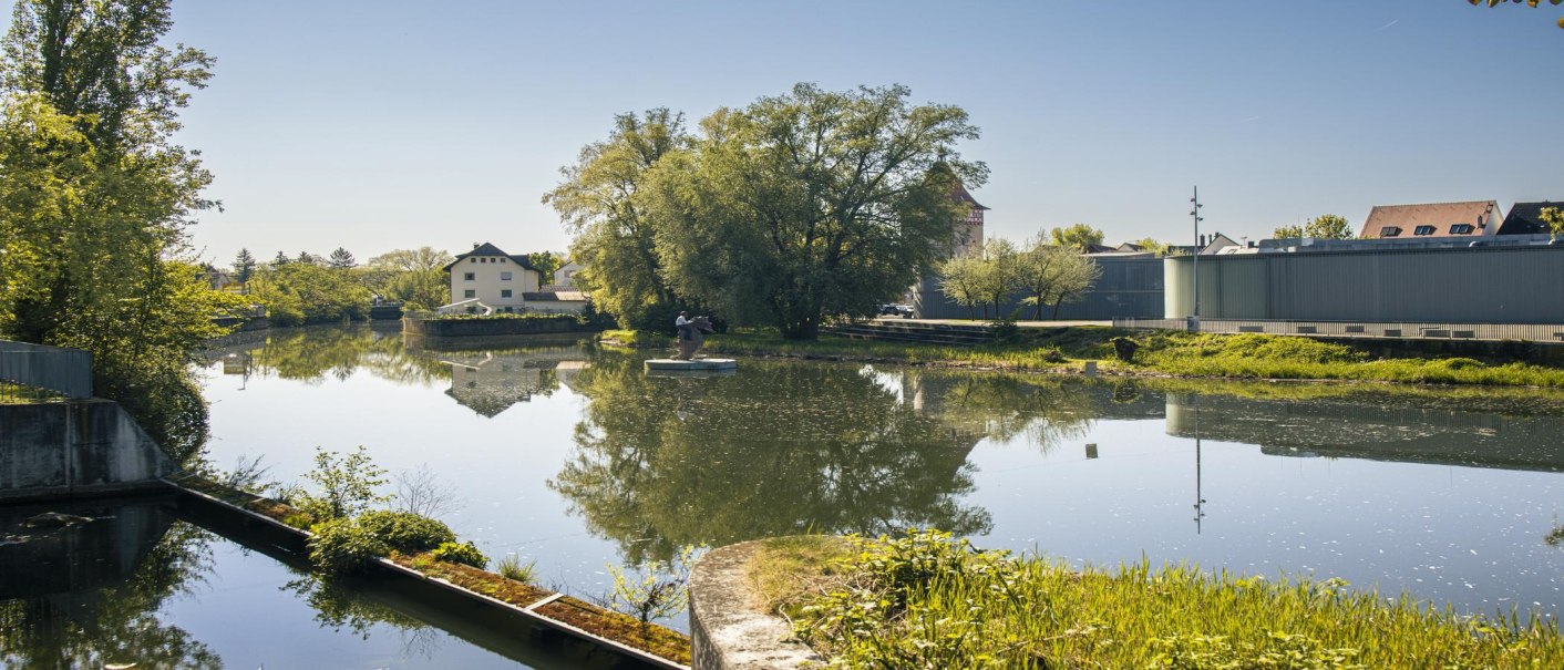 A calm river with trees and buildings on the banks, including the Stihl Gallery in Waiblingen. Clear reflection in the water under a blue sky., © SMG Stuttgart Marketing GmbH - Sarah Schmid