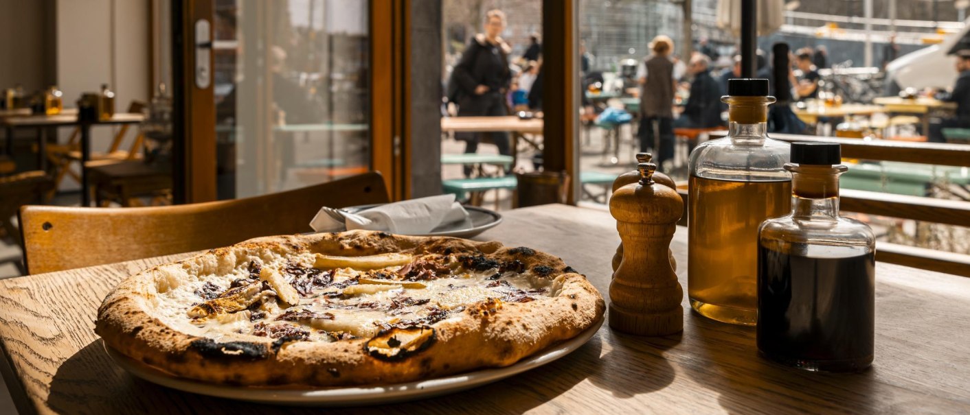 Eine Pizza auf einem Holztisch in einem Restaurant. Im Hintergrund sind Menschen auf einer Terrasse zu sehen. Auf dem Tisch stehen Ölflaschen und ein Pfefferstreuer., © SMG, Sarah Schmid Eine Pizza auf einem Holztisch in einem Restaurant. Im Hintergrund sind Menschen auf einer Terrasse zu sehen. Auf dem Tisch stehen Ölflaschen und ein Pfefferstreuer., © SMG, Sarah Schmid