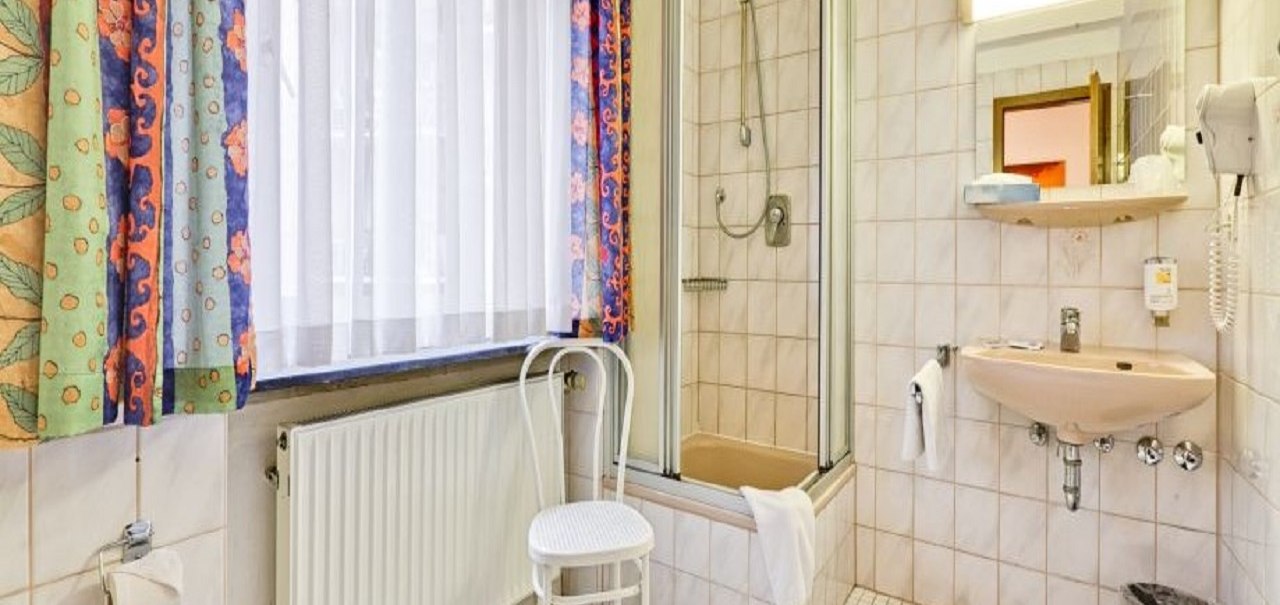 Ein helles Badezimmer mit Dusche, Waschbecken, Stuhl und bunten Vorhängen. Fliesen an Wänden und Boden, Handtücher und ein Föhn sind vorhanden., © ARNOTEL GmbH