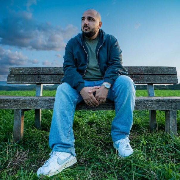 Ein Mann sitzt auf einer Holzbank in einer Wiese, trägt Jeans und eine Jacke. Der Himmel ist blau mit Wolken., © SBEntertainment GmbH & Co. KG Ein Mann sitzt auf einer Holzbank in einer Wiese, trägt Jeans und eine Jacke. Der Himmel ist blau mit Wolken., © SBEntertainment GmbH & Co. KG