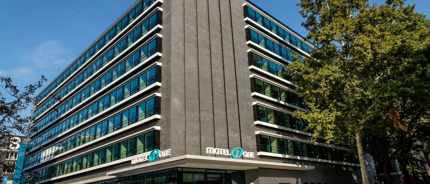 Modern building with glass facade and Motel One logo, surrounded by trees, under a blue sky., &copy; Motel One - Stuttgart Hauptbahnhof