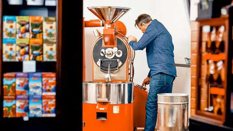 Ein Mann in Jeans bedient eine große, orangefarbene Kaffeeröstmaschine. Im Hintergrund sind bunte Kaffeepackungen zu sehen., © Kaffee Fröhlich, Stuttgart