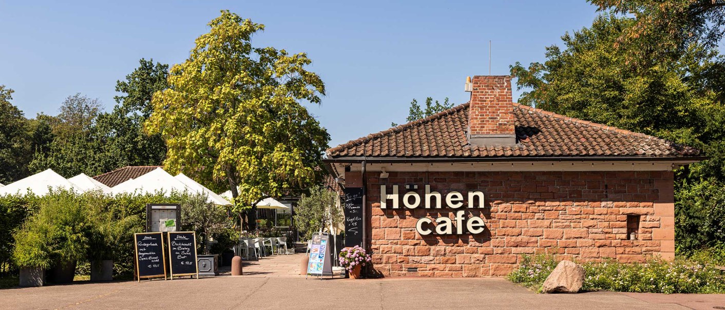 The Höhencafé in Killesbergpark is a brick building with a terrace surrounded by trees and plants. White parasols are visible., © Stuttgart-Marketing GmbH, Sarah Schmid The Höhencafé in Killesbergpark is a brick building with a terrace surrounded by trees and plants. White parasols are visible., © Stuttgart-Marketing GmbH, Sarah Schmid