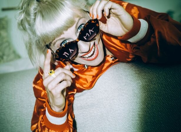 Frau mit blonden Haaren und Sonnenbrille, die "MAMA" zeigt, lächelt und trägt eine orange Jacke., © Theaterhaus Stuttgart e.V. Frau mit blonden Haaren und Sonnenbrille, die "MAMA" zeigt, lächelt und trägt eine orange Jacke., © Theaterhaus Stuttgart e.V.