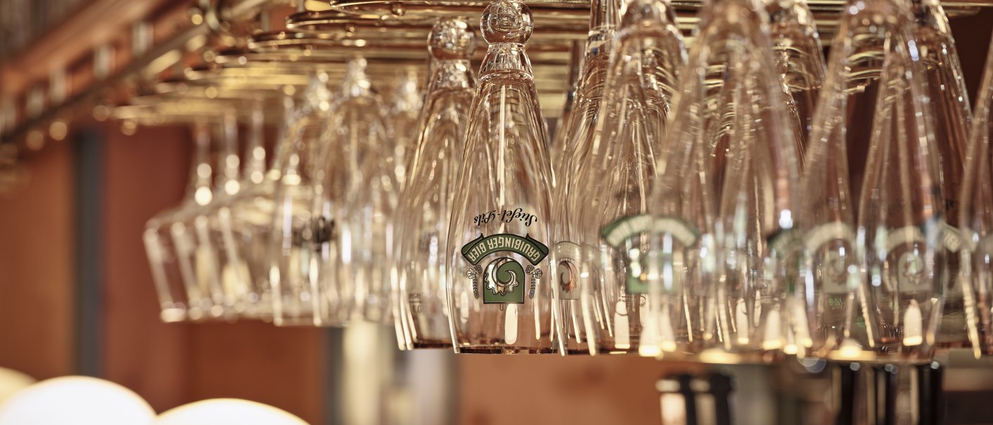 Hängende Biergläser in einer Bar, warm beleuchtet. Die Gläser sind kopfüber an einem Gestell befestigt, im Hintergrund sind Lampen zu sehen., © Badhotel Restaurant Stauferland