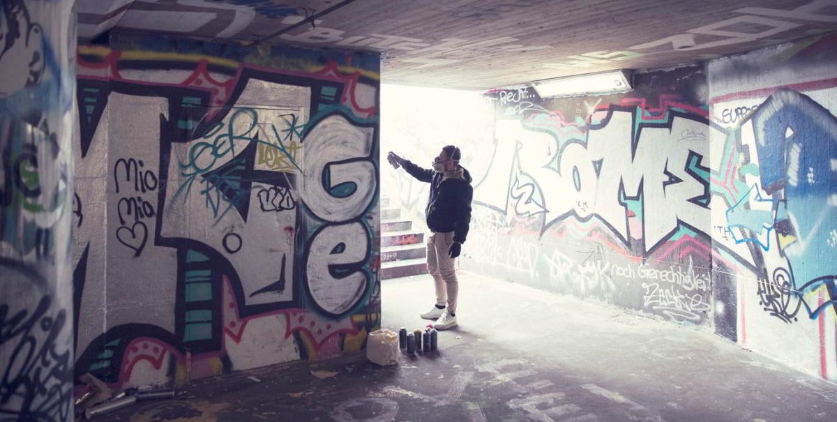 Eine Person sprüht Graffiti in einem Tunnel. Die Wände sind mit bunten Graffitis bedeckt. Auf dem Boden stehen Farbdosen., © hoponopo GmbH Eine Person sprüht Graffiti in einem Tunnel. Die Wände sind mit bunten Graffitis bedeckt. Auf dem Boden stehen Farbdosen., © hoponopo GmbH