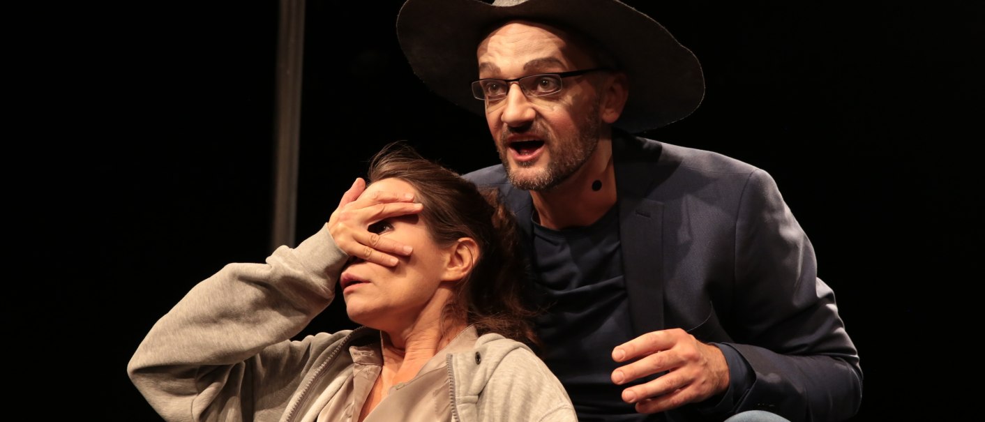 Ein Mann mit Hut und Brille spricht auf der Bühne, während er die Augen einer Frau verdeckt. Beide tragen Mikrofone., © Theater tri-bühne, Stefan Kirchknopf Ein Mann mit Hut und Brille spricht auf der Bühne, während er die Augen einer Frau verdeckt. Beide tragen Mikrofone., © Theater tri-bühne, Stefan Kirchknopf