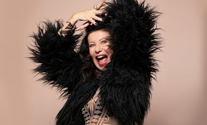 Eine Frau in einem schwarzen, flauschigen Outfit posiert fr&ouml;hlich vor einem beigen Hintergrund. Sie lacht und hebt die Arme in einer dynamischen Pose., &copy; Theaterhaus Stuttgart e.V.