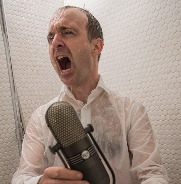 Ein Mann in einem wei&szlig;en Hemd singt leidenschaftlich in einer Dusche, w&auml;hrend er ein Mikrofon h&auml;lt. Wasser str&ouml;mt &uuml;ber ihn., &copy; Rosenau Kultur e.V.