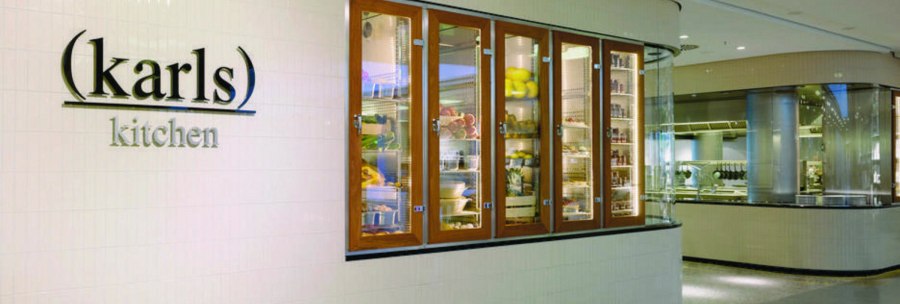 Entrance to "karls kitchen" with illuminated glass cabinets full of food and a view of the modern kitchen., © karls kitchen, Stuttgart