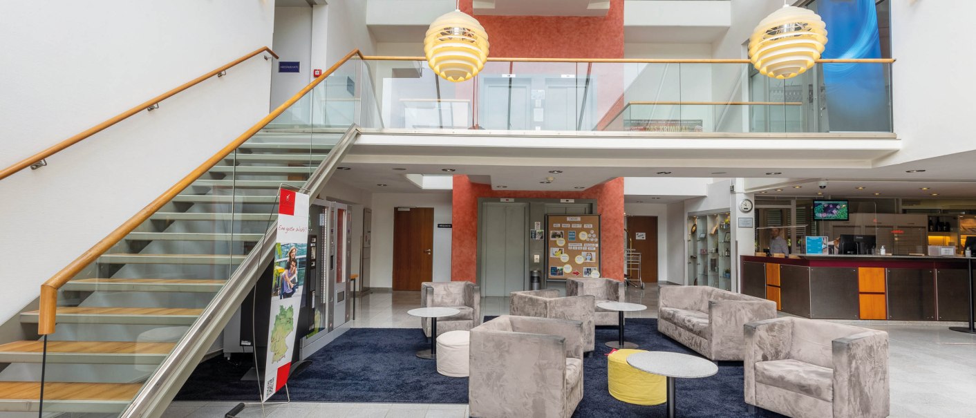 Moderne Lobby mit Sitzbereich, grauen Sesseln, Treppe mit Glasgeländer und Rezeption. Helle Beleuchtung und offene Architektur., © Seminaris Hotel Bad Boll