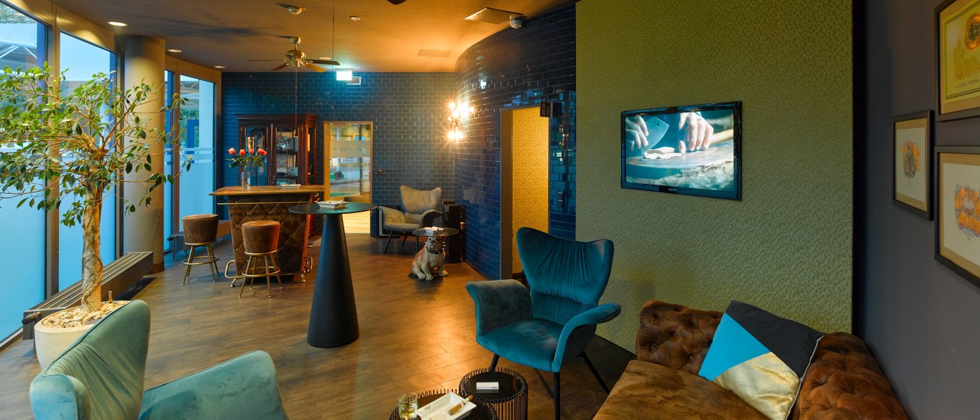 Elegant cigar lounge with blue armchairs, brown sofa, bar area and stylish decoration. A television shows a close-up of hands., © Holiday Inn Stuttgart Elegant cigar lounge with blue armchairs, brown sofa, bar area and stylish decoration. A television shows a close-up of hands., © Holiday Inn Stuttgart