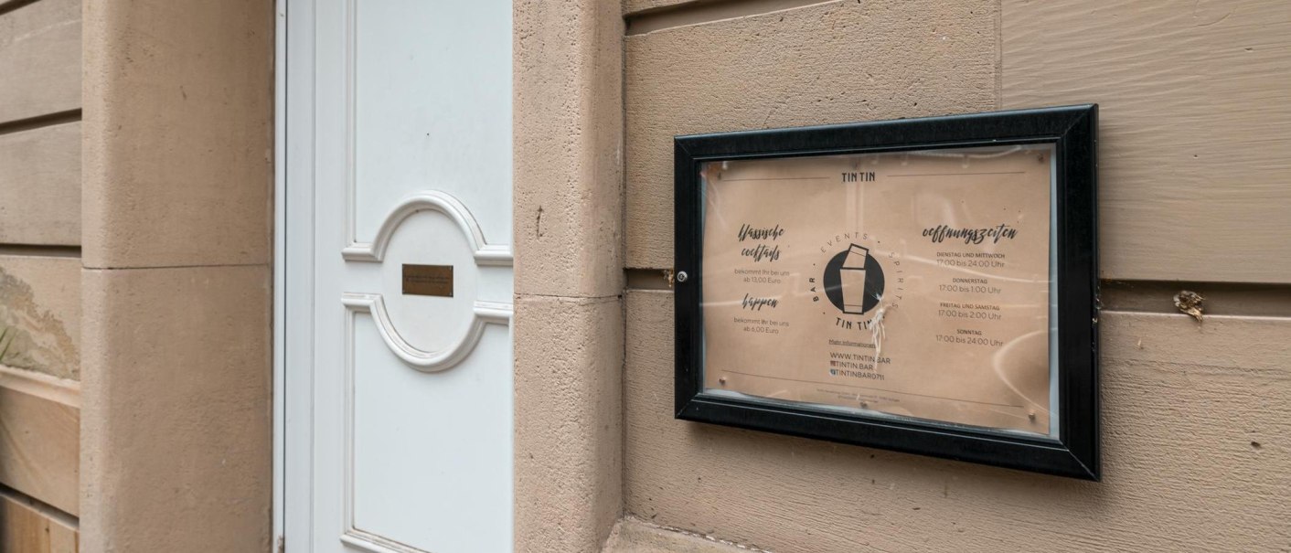 Schild der TinTin Bar mit Öffnungszeiten und Preisen an einer Wand neben einer weißen Tür., © SMG, Sarah Schmid