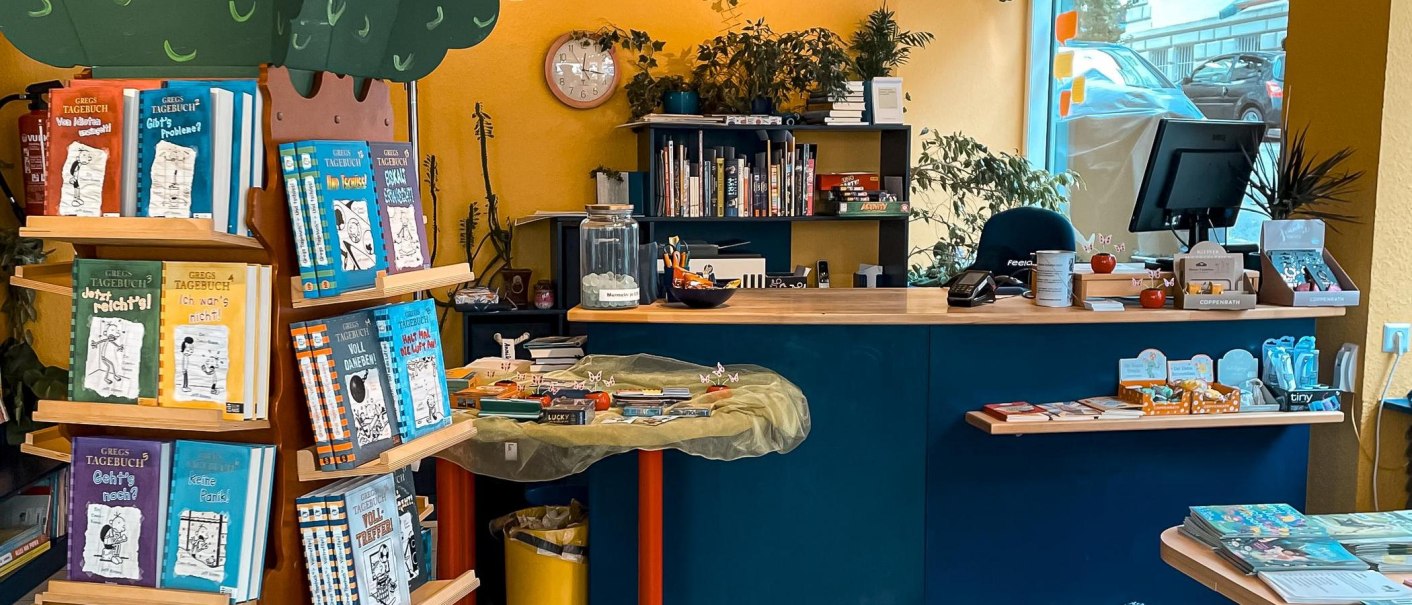 Ein bunter Buchladen mit Regalen voller Bücher, darunter "Gregs Tagebuch". Ein Verkaufstresen mit Computer und Dekorationen ist zu sehen., © Naseweis, Stuttgart