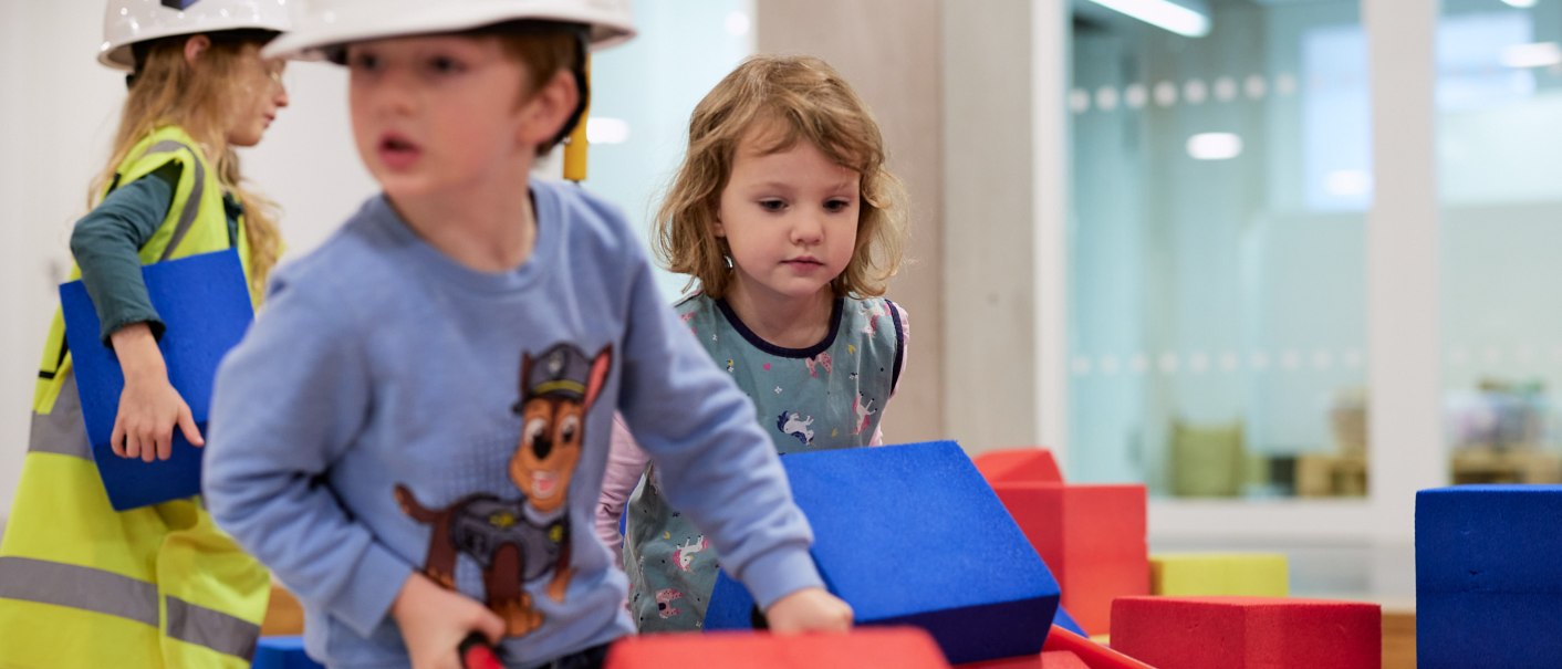 Kinder spielen auf einer Indoor-Baustelle mit bunten Bauklötzen. Sie tragen Bauhelme und Sicherheitswesten. Im Hintergrund sind moderne Räume zu sehen., © Julia Ochs