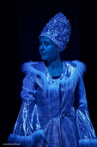 Eine Frau in einem glitzernden Kostüm mit Fellbesatz und einer Krone, beleuchtet von blauem Licht, stellt die Schneekönigin dar., © Theater Atelier