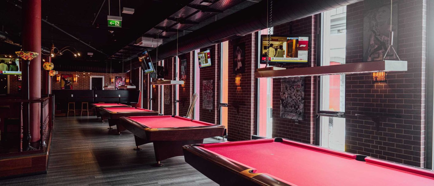 Ein stilvoller Billardraum mit roten Tischen, mehreren Fernsehern und Backsteinw&auml;nden. Die Beleuchtung ist ged&auml;mpft und gem&uuml;tlich., &copy; Palm Beach Stuttgart GmbH