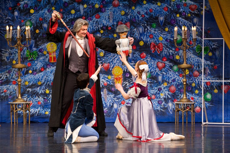 Ballettszene aus dem Nussknacker: Ein Mann in rotem Mantel hält eine Nussknackerfigur, während zwei Tänzer in historischen Kostümen knien. Hintergrund: Weihnachtsbaum., © Go 2 - Convent GmbH Ballettszene aus dem Nussknacker: Ein Mann in rotem Mantel hält eine Nussknackerfigur, während zwei Tänzer in historischen Kostümen knien. Hintergrund: Weihnachtsbaum., © Go 2 - Convent GmbH