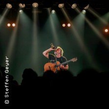 Ein Musiker steht auf einer Bühne, spielt Gitarre und singt in ein Mikrofon. Die Bühne ist von Scheinwerfern beleuchtet, das Publikum ist als Silhouette zu sehen., © links im Bild Ein Musiker steht auf einer Bühne, spielt Gitarre und singt in ein Mikrofon. Die Bühne ist von Scheinwerfern beleuchtet, das Publikum ist als Silhouette zu sehen., © links im Bild