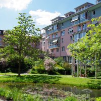 Mehrstöckiges Wohngebäude mit rosa und blauen Fassaden, umgeben von einem grünen Garten mit Bäumen und einem kleinen Teich., © Ev. Diakonissenanstalt Stuttgart Mehrstöckiges Wohngebäude mit rosa und blauen Fassaden, umgeben von einem grünen Garten mit Bäumen und einem kleinen Teich., © Ev. Diakonissenanstalt Stuttgart