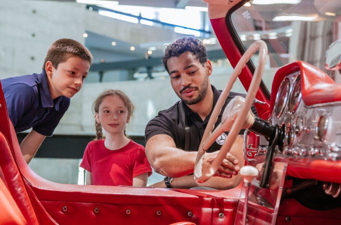 A man explains the cockpit of a red vintage car in a modern museum to two children. The children watch with interest., &copy; Mercedes-Benz Heritage GmbH