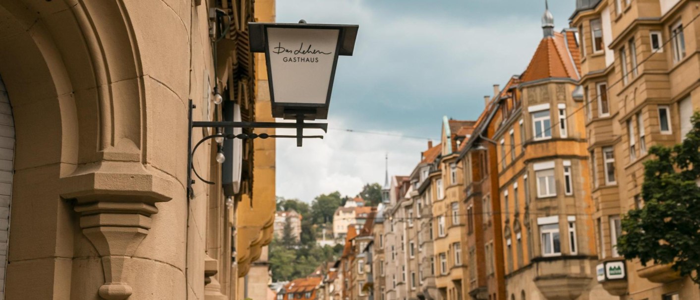 Straßenszene in Stuttgart mit einem Gasthausschild "Das Lehen" und historischen Gebäuden im Hintergrund., © SMG, Sarah Schmid