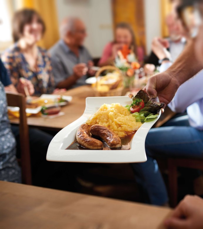 Ein Teller mit geschmorten Bratw&uuml;rsten, Kartoffelp&uuml;ree und Salat wird in einem gem&uuml;tlichen Restaurant serviert. Im Hintergrund sitzen Menschen an einem Holztisch und genie&szlig;en ihr Essen in geselliger Runde., &copy; SMG, Jean-Claude Winkler