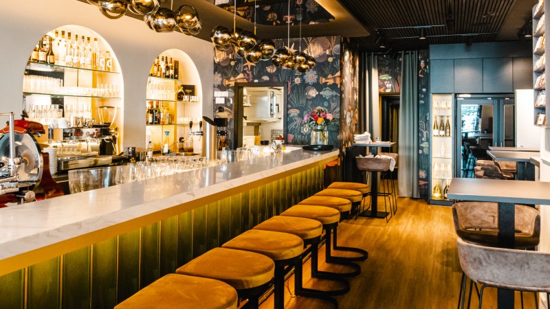 Elegante Bar mit langer Theke, gelben Barhockern und stilvollen Lampen. Die Wände sind mit floralen Mustern dekoriert, Flaschenregale im Hintergrund., © SMG, Sarah Schmid Elegante Bar mit langer Theke, gelben Barhockern und stilvollen Lampen. Die Wände sind mit floralen Mustern dekoriert, Flaschenregale im Hintergrund., © SMG, Sarah Schmid