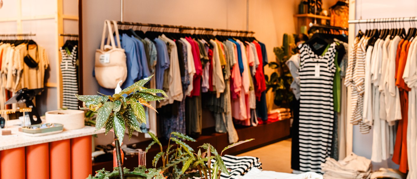 Ein Bekleidungsgeschäft mit bunten Kleidern auf Kleiderstangen, Pflanzen und einem Spiegel im Hintergrund., © SMG_Sarah Schmid Ein Bekleidungsgeschäft mit bunten Kleidern auf Kleiderstangen, Pflanzen und einem Spiegel im Hintergrund., © SMG_Sarah Schmid