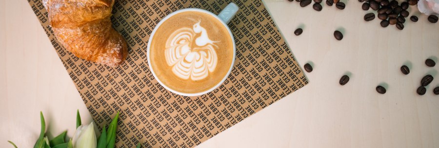 A croissant and cappuccino with latte art on a table surrounded by coffee beans, cotton, roses and tulips., © CELBRE A croissant and cappuccino with latte art on a table surrounded by coffee beans, cotton, roses and tulips., © CELBRE