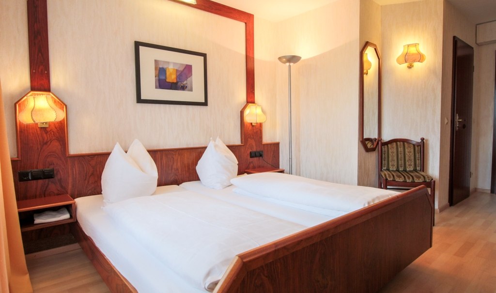 Ein komfortables Doppelzimmer mit Holzbetten, zwei Nachttischlampen, einem Kunstwerk an der Wand und einem Stuhl in der Ecke., © Ulli Kellenbenz