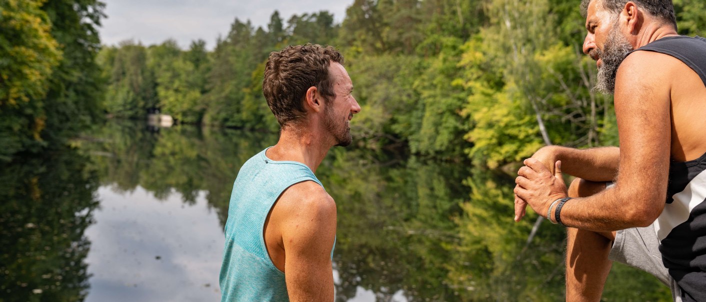 Two men sit on the shore of a lake, surrounded by green trees, and talk. One of them is holding a water bottle., &copy; SMG, Martina Denker