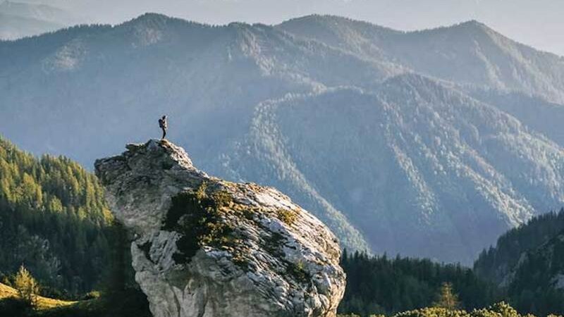 A person stands on a large rock, surrounded by an impressive mountain landscape with wooded hills and snow-capped peaks., &copy; Theaterhaus Stuttgart e.V.